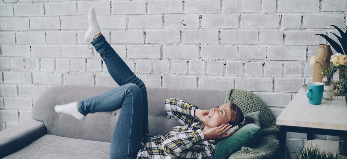 Joyful young woman is singing, listening to music in headphones and moving hands and legs dancing lying on sofa in modern loft style apartment. People, technology and fun concept.