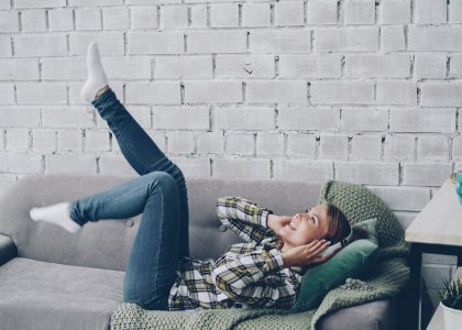 Joyful young woman is singing, listening to music in headphones and moving hands and legs dancing lying on sofa in modern loft style apartment. People, technology and fun concept.