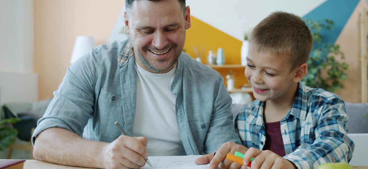 Cheerful dad is drawing funny pictures for happy child having fun sitting at desk at home enjoying leisure time indoors. Youth, creative people and arts concept.
