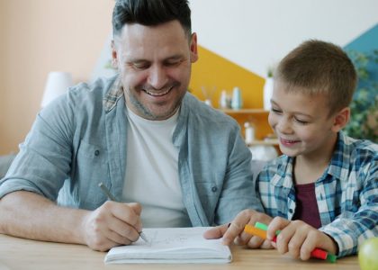 Cheerful dad is drawing funny pictures for happy child having fun sitting at desk at home enjoying leisure time indoors. Youth, creative people and arts concept.